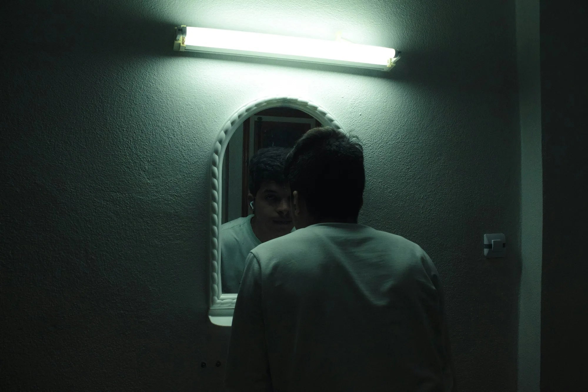 A man standing quietly in front of a bathroom mirror, a reflective moment evoking the kind of self-examination and worry behind common hair loss myths