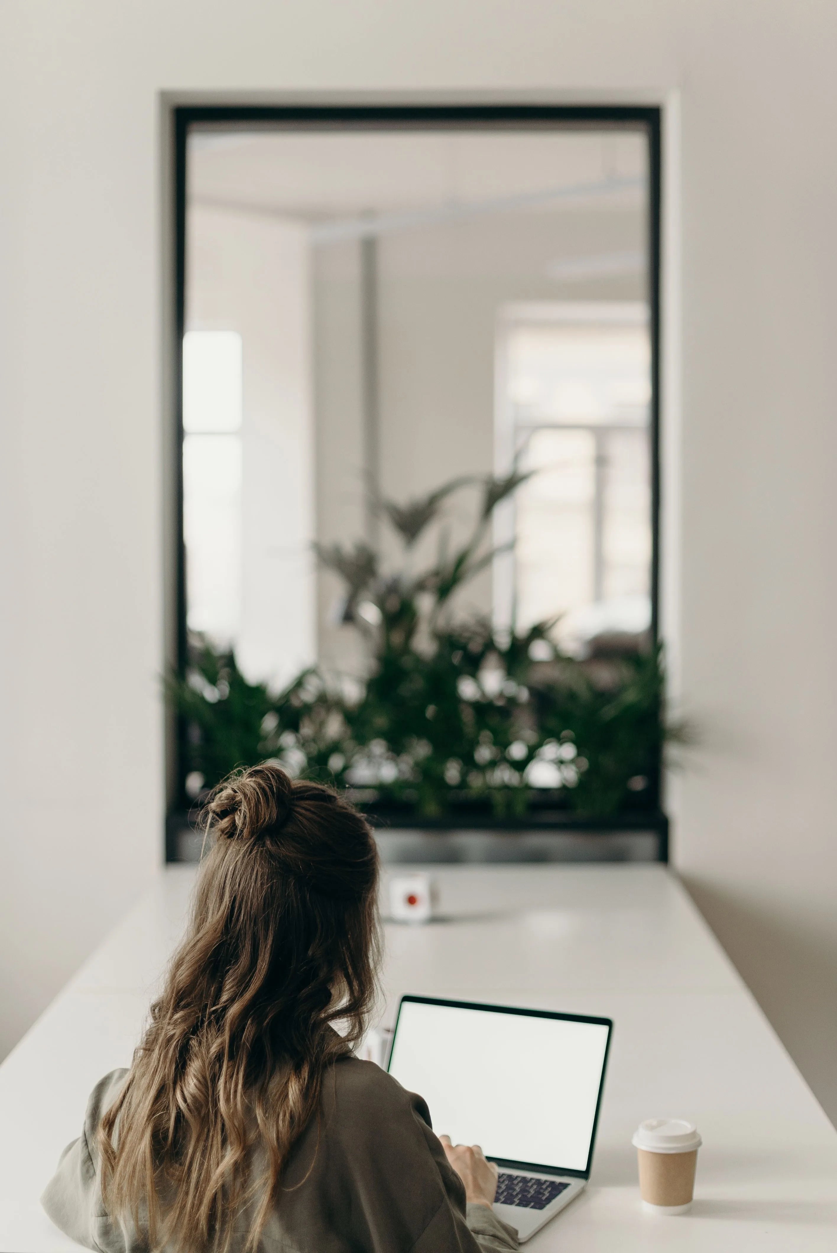 Woman working on a laptop at a bright, minimalist desk, representing the choice between an online telehealth consultation and an at home hair care routine