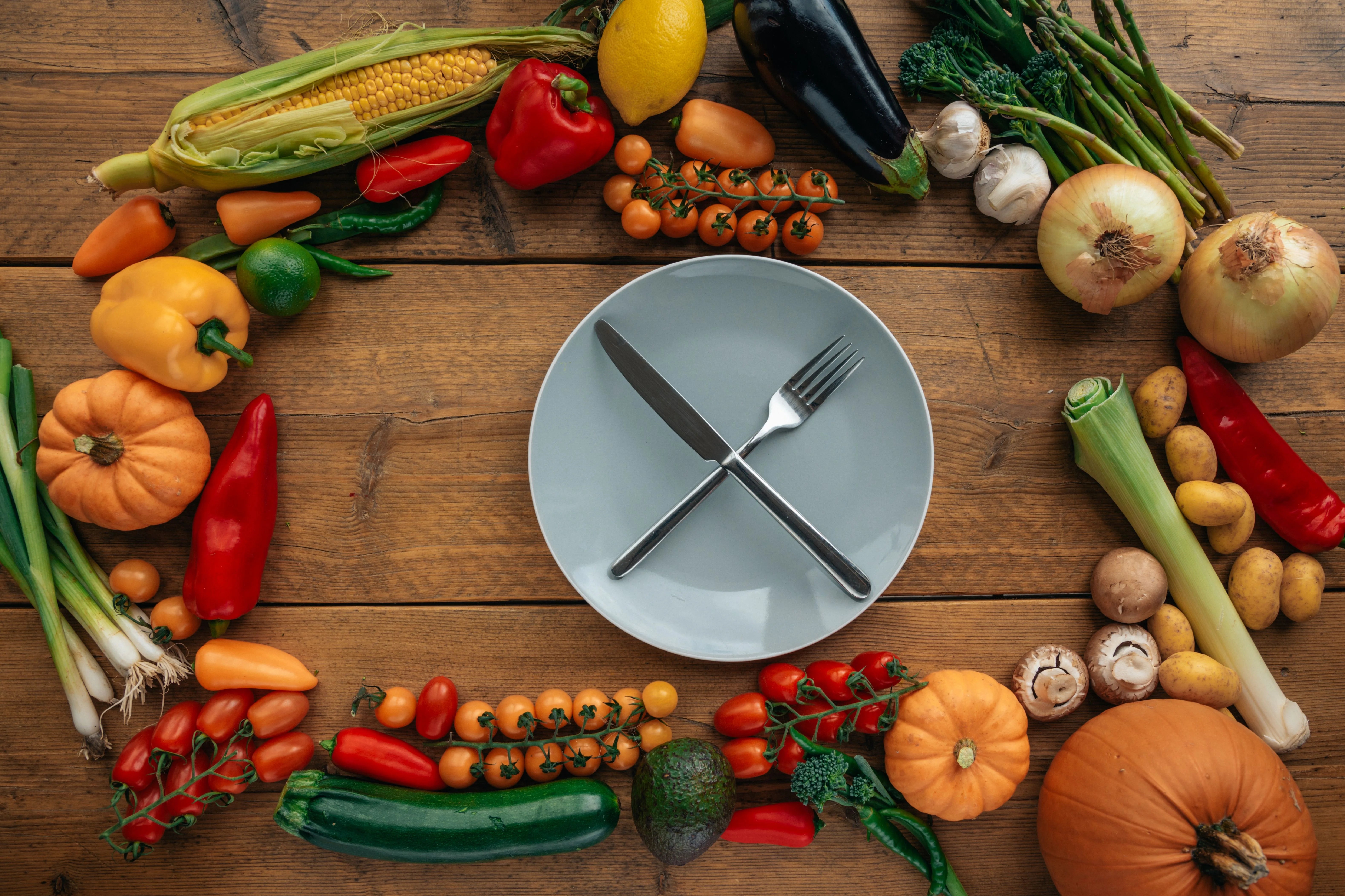 Overhead flat lay of an empty ceramic plate surrounded by fresh, colorful vegetables on a wooden table, representing mindful eating and meal timing