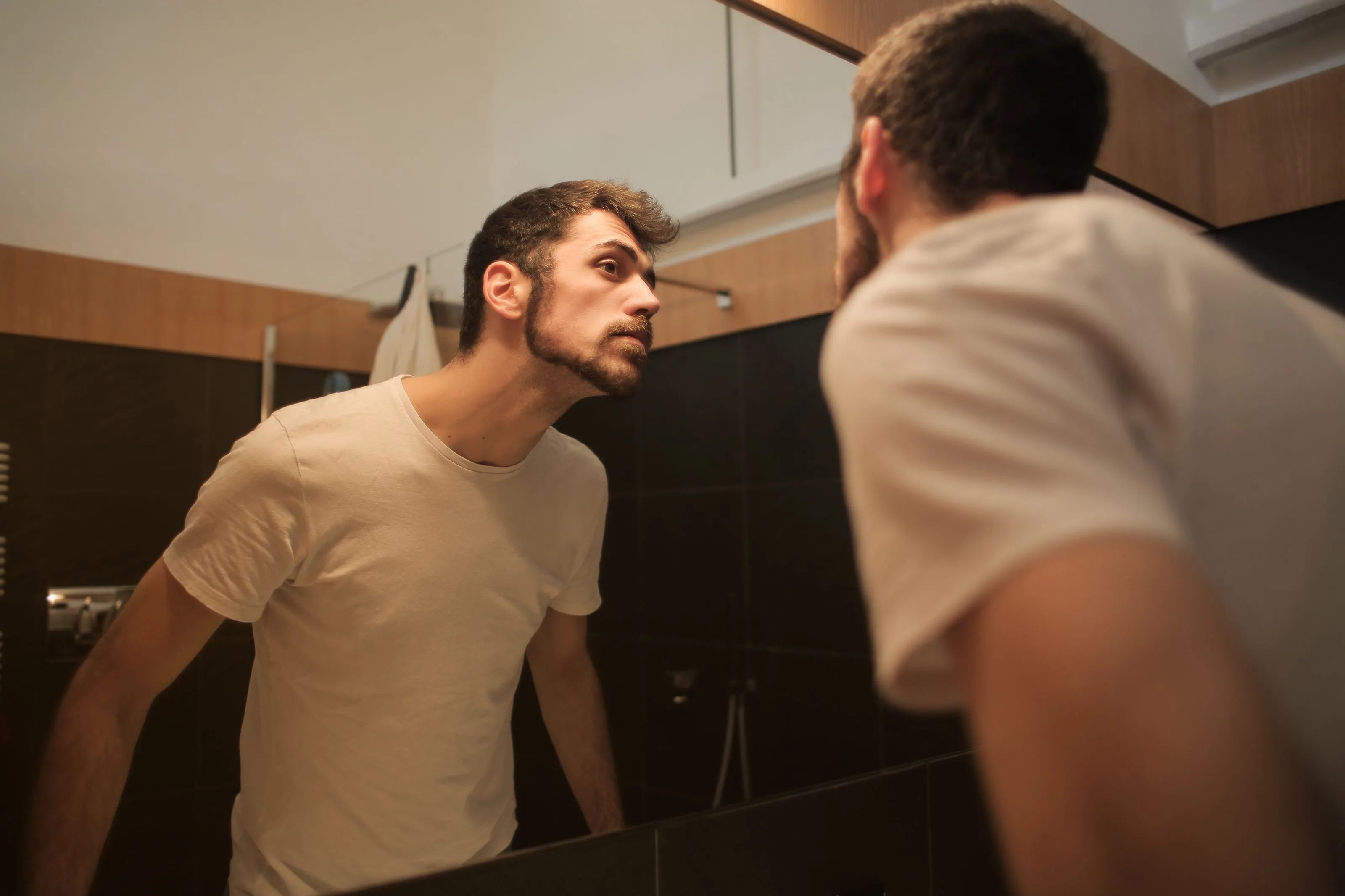 Man closely examining his hair and hairline in a bathroom mirror while considering hair treatment options