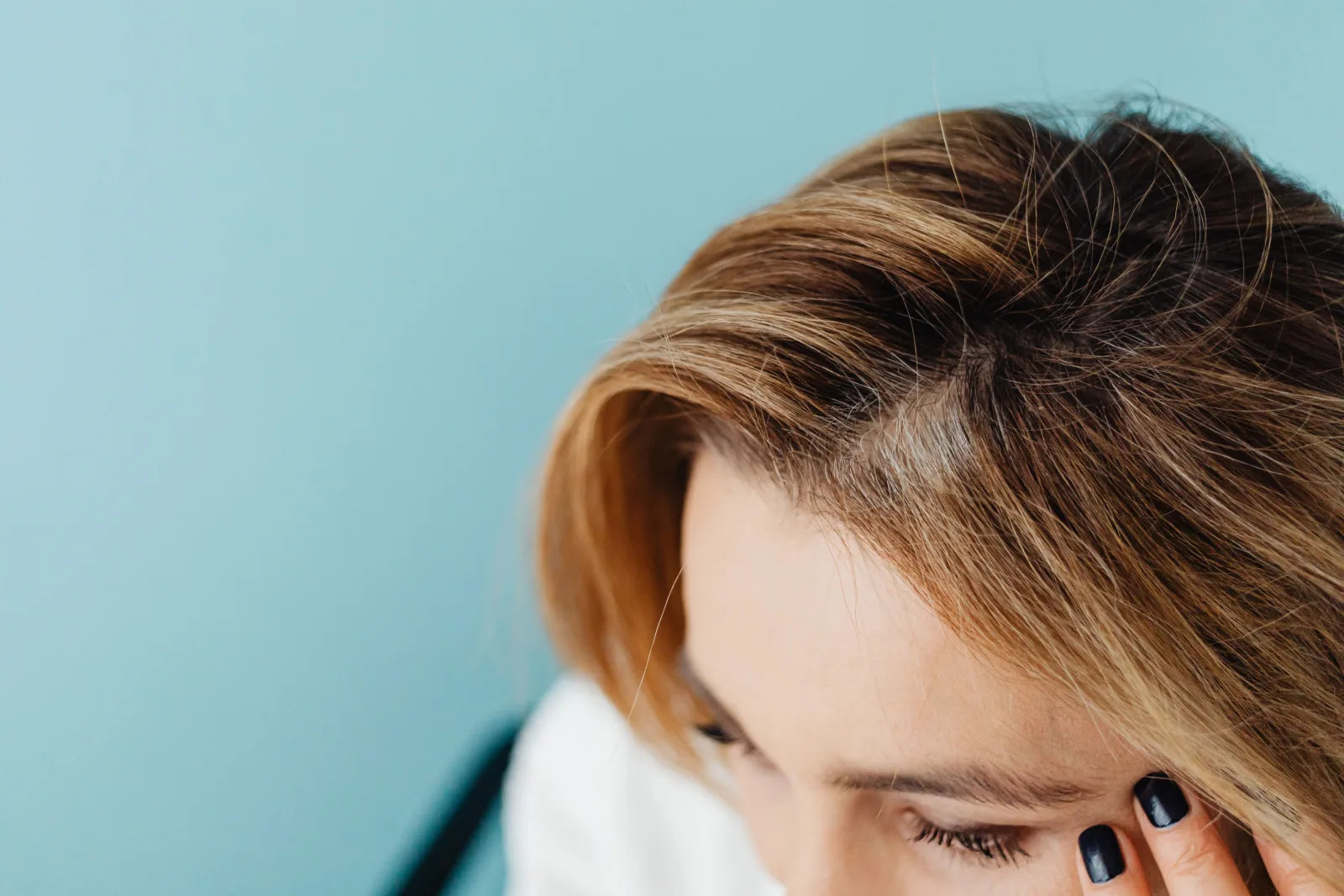 Close-up of a woman gently touching her forehead and hairline, showing her natural hair part against a soft blue background