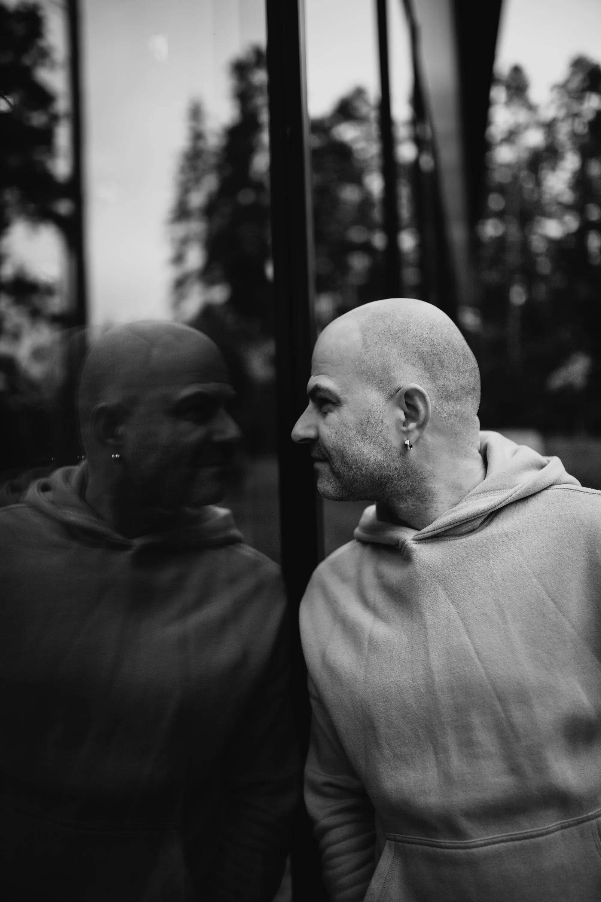A bald man in a dark hoodie looking thoughtfully at his own reflection, symbolizing the personal decision-making involved in choosing between a telehealth hair loss prescription and a non-prescription hair regrowth system